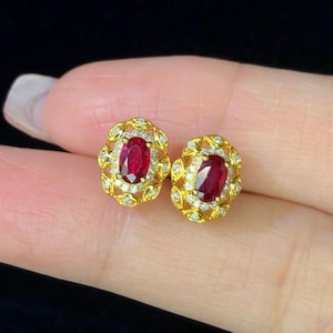 Może przedstawiać: Para złotych kolczyków sztyftów. Każdy kolczyk ma duży, owalny, ciemnoczerwony kamień szlachetny, otoczony mniejszymi, przezroczystymi kamieniami szlachetnymi i ozdobną, złotą filigranową obwódką. Neutralne tło.