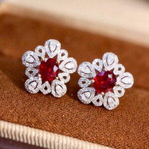 Może przedstawiać: Para kolczyków w kształcie kwiatów. Każdy kolczyk ma w środku żywy, czerwony owalny kamień szlachetny, otoczony aureolą mniejszych, błyszczących białych kamieni. Kolczyki osadzone są na złotej podstawie.