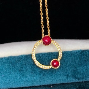 May include: A gold necklace with a circular pendant. The pendant has a ring of small, clear stones and two round, red gemstones. The necklace chain is gold-coloured and delicate.