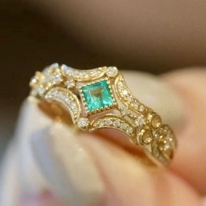 May include: A gold ring with a square emerald gemstone surrounded by smaller diamonds. The ring has a delicate, intricate design with a filigree pattern.