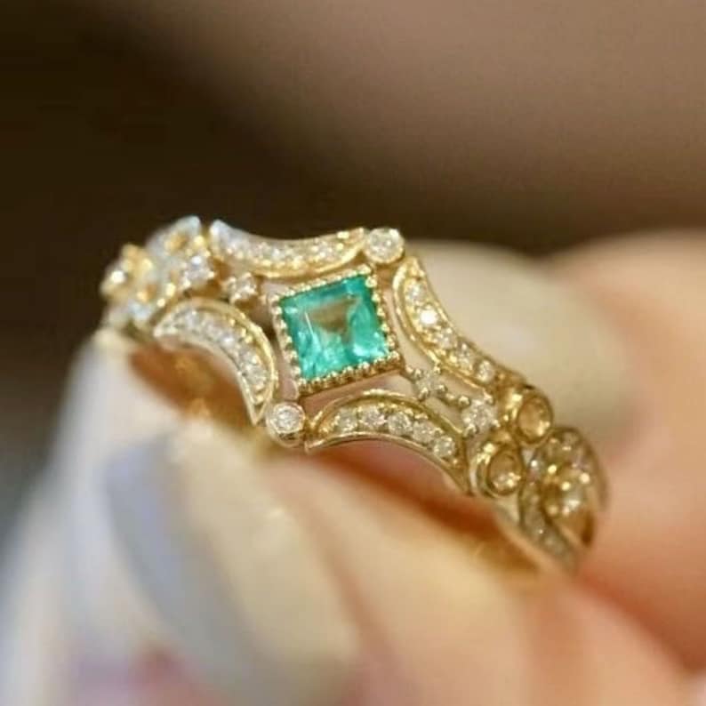 May include: A gold ring with a square emerald gemstone surrounded by smaller diamonds. The ring has a delicate, intricate design with a filigree pattern.