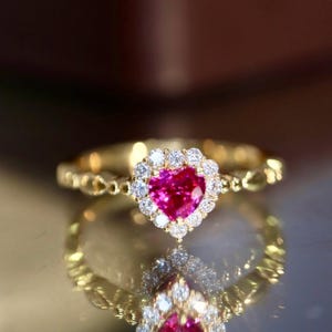 Peut inclure: Une bague en or ornée d'une pierre précieuse rose vif en forme de cœur, entourée d'un halo de petites pierres claires. La monture de la bague présente un motif texturé et délicat. La bague est présentée sur une surface réfléchissante.
