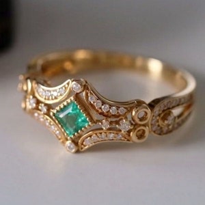 May include: A gold ring with a square emerald gemstone surrounded by smaller diamonds. The ring has a delicate, intricate design with a filigree pattern.