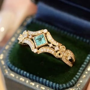 May include: A gold ring with a square emerald gemstone surrounded by smaller diamonds. The ring has a delicate, intricate design.