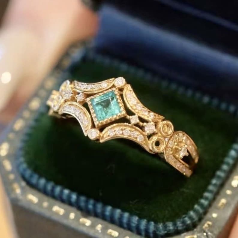 May include: A gold ring with a square emerald gemstone surrounded by smaller diamonds. The ring has a delicate, intricate design.