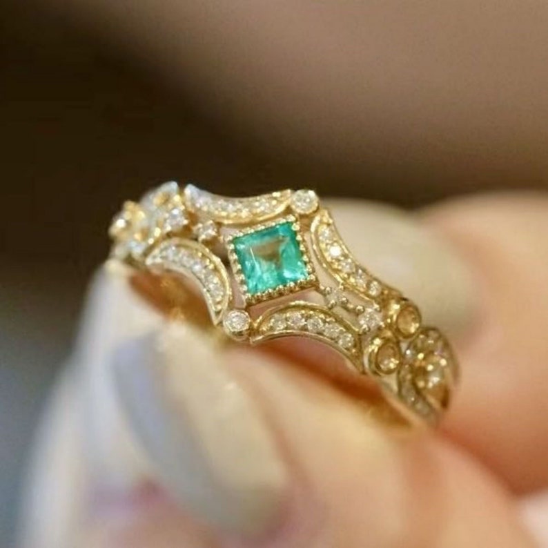 May include: A gold ring with a square emerald gemstone surrounded by smaller diamonds. The ring has a delicate, intricate design.