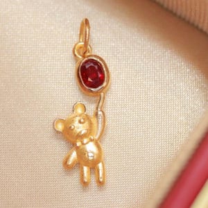 May include: A gold-coloured pendant featuring a teddy bear holding a red gemstone balloon. The teddy bear has a bow tie and is holding the balloon with its right hand. The gemstone is oval-shaped.