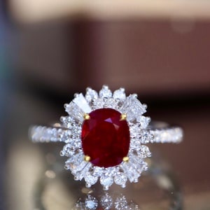 Könnte beinhalten: Nahaufnahme eines Rings mit einem großen, oval geschliffenen Rubin-Edelstein, umgeben von einem Halo aus kleineren, klaren Diamanten. Das Band des Rings ist ebenfalls mit Diamanten besetzt, was einen funkelnden Effekt erzeugt.