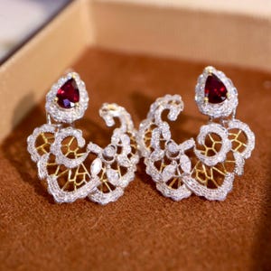 May include: A pair of elaborate earrings with a teardrop-shaped, deep red gemstone at the top, encircled by small, sparkling stones. The earrings have a gold filigree design with additional clear stones.
