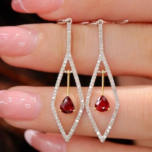 May include: A pair of diamond and ruby earrings. Each earring features a teardrop-shaped, deep red ruby suspended from a gold bar within a diamond-encrusted, elongated diamond-shaped frame. The earrings are held up by a silver hook.