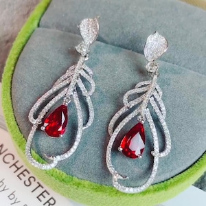 May include: A pair of drop earrings with a leaf design. Each earring has a pear-shaped, deep red gemstone hanging from a silver-coloured, leaf-like structure, embellished with small, sparkling stones. The earrings are displayed on a grey surface.