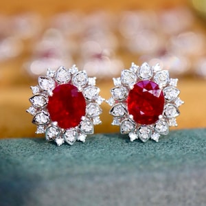 以下が含まれることがあります： 大きな楕円形の深い赤色の宝石を、小さな透明な宝石のハローで囲んだスタッドピアス。 イヤリングは銀色の金属にセットされ、緑色の表面に置かれています。