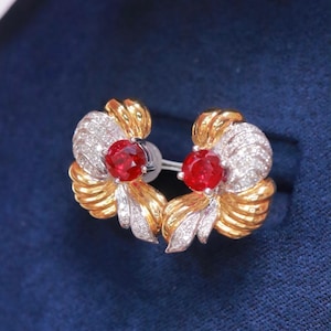 May include: A pair of stud earrings featuring a vibrant red gemstone center, surrounded by a gold and silver floral design. The earrings have a post backing and are set against a dark blue background.