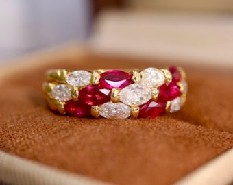 Puede incluir: Un anillo de oro con filas alternas de piedras preciosas rojas y blancas. Las piedras preciosas, de forma ovalada, están engastadas juntas, creando una banda continua de color. El anillo se muestra sobre una superficie marrón.