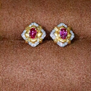 May include: A pair of stud earrings featuring a central oval-cut pink gemstone surrounded by a textured gold frame and an outer border of small clear stones. The design is symmetrical with a floral-inspired shape and a polished metallic finish.
