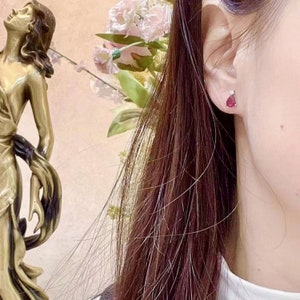 Pu&ograve; includere: Primo piano di un orecchio con un piccolo orecchino a forma di lacrima di pietra preziosa rossa. L'orecchino &egrave; incastonato in una montatura d'argento o d'oro bianco.
