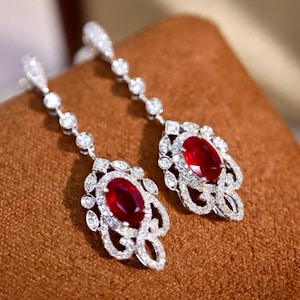 May include: A pair of drop earrings with oval red gemstones and surrounding white stones. The earrings have an intricate design with smaller stones leading up to the ear wire. The jewellery is displayed on a brown surface.