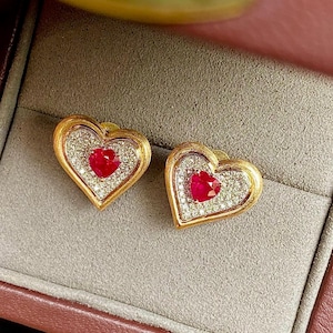 以下が含まれることがあります： ハート型のイヤリングのペア。各イヤリングは、小さな透明な石のハローに囲まれた中央の赤い宝石を特徴としています。外側の縁は金色です。イヤリングはジュエリーボックスに入っています。