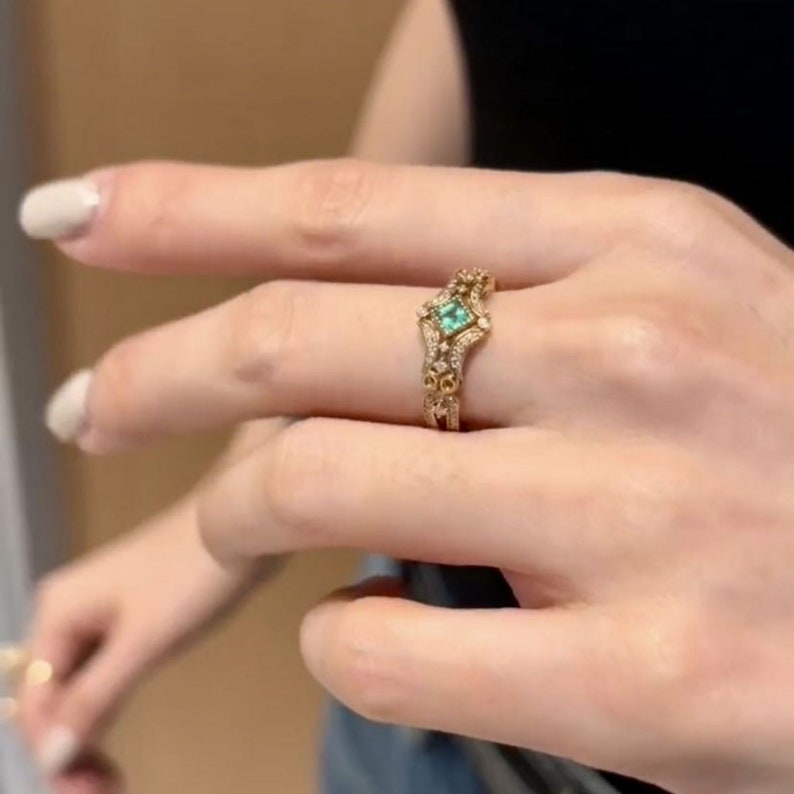 May include: A gold ring with a square emerald green gemstone surrounded by smaller diamonds. The ring has a delicate, intricate design.