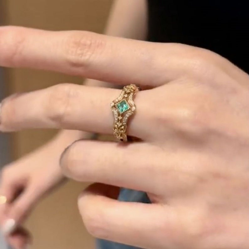 May include: A gold ring with a green gemstone set in a square shape. The ring is adorned with intricate details and smaller gemstones.