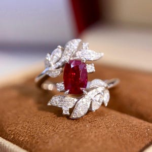 May include: A silver ring with a large, rectangular, deep red gemstone at its centre. The ring is decorated with leaf-like designs encrusted with small, sparkling white gemstones. The ring is set against a blurred brown background.