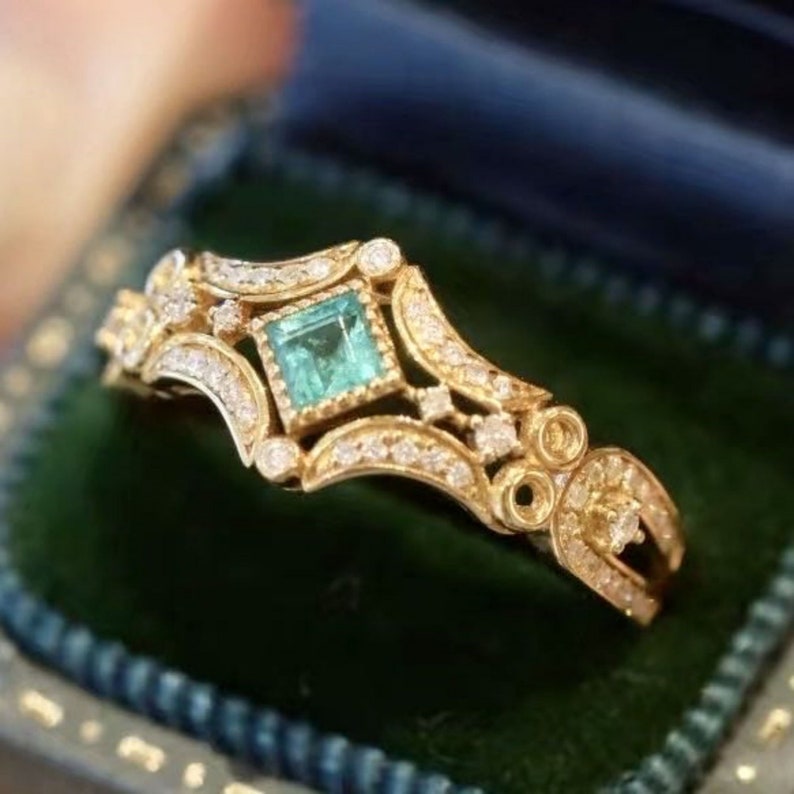 May include: A gold ring with a square emerald green gemstone surrounded by smaller diamonds. The ring has a delicate, intricate design.