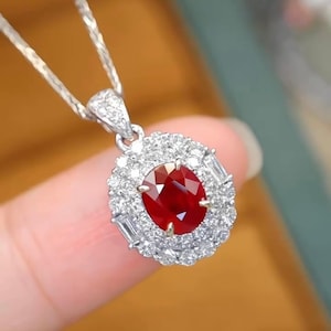 May include: A silver necklace with a pendant featuring a large, oval-cut ruby surrounded by a halo of small, round diamonds. The pendant also has several baguette-cut diamonds. The ruby is a deep red color.