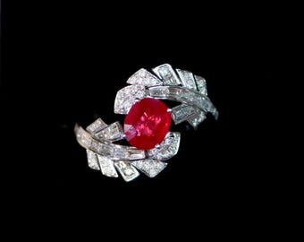 Puede incluir: Un anillo de plata con una gran gema de rubí de corte ovalado. El diseño del anillo incorpora dos elementos estilizados en forma de hoja, incrustados con diamantes más pequeños y brillantes. El anillo está sobre un fondo negro.