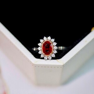 May include: A gemstone ring featuring a large oval-cut, deep red ruby center stone. The ruby is surrounded by a halo of small, round, white diamonds. The ring has a silver band with gold accents.