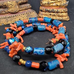 May include: A layered necklace featuring coral-colored, blue, and black beads. The necklace incorporates cylindrical blue beads with a white swirl design, interspersed with coral-colored branch-like beads and black faceted beads. Gold-colored beads add accents.