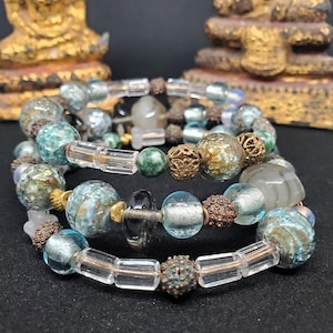 May include: A set of four bracelets featuring a mix of clear, green, and blue glass beads, along with gold and silver accents. The bracelets are stacked on top of each other, creating a layered look.