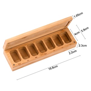 K&ouml;nnte beinhalten: Ein rechteckiger Pillen-Organizer aus Bambus mit Scharnierdeckel. Die offene Box zeigt sieben F&auml;cher, die f&uuml;r jeden Wochentag beschriftet sind. Die Box ist 19,8 cm lang, 2,3 cm hoch und 5,9 cm breit. Der Deckel ist 1,05 cm dick.