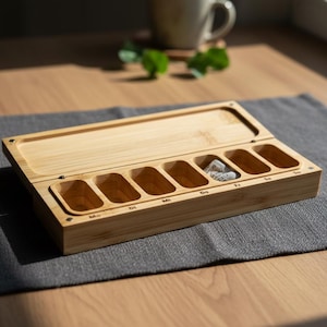 K&ouml;nnte beinhalten: Ein rechteckiger Pillen-Organizer aus Bambus mit Klappdeckel. Der Organizer hat sieben F&auml;cher, die mit den Wochentagen beschriftet sind und jeweils zur Aufnahme von Pillen dienen. Die Box ist aus hellem Holz gefertigt und liegt auf einem grauen Platzset.