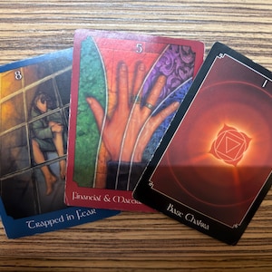 Könnte beinhalten: Drei Tarotkarten liegen auf einer Holzoberfläche. Die Karten zeigen Illustrationen einer Person hinter Gittern, einer Hand und eines geometrischen Designs. Auf den Karten steht "Trapped in Fear", "Financial & Macro" und "Base Chakra".
