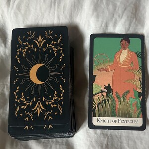 Könnte beinhalten: Eine Tarotkarte mit schwarzem Hintergrund und goldenen Akzenten. Die Karte zeigt einen zunehmenden Mond und eine Sonne mit einem floralen Design. Die Karte ist mit "Knight of Pentacles" beschriftet.