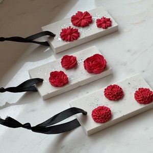 May include: Three white rectangular ceramic air fresheners with red flower designs and black ribbon hangers. The air fresheners are on a white marble surface.