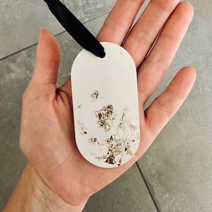 Könnte beinhalten: Ein ovales, weißes Etikett mit Goldblatt-Akzenten und einem schwarzen Band. Das Etikett wird in einer Hand gehalten, wobei die Goldblattdetails hervorgehoben werden. Es handelt sich um einen dekorativen Artikel.