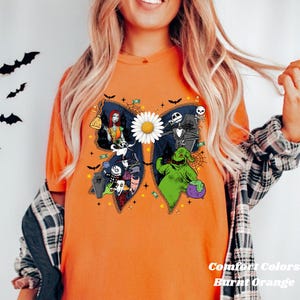 May include: Burnt orange t-shirt with a large butterfly design featuring characters from The Nightmare Before Christmas. The butterfly has a white daisy centre and is surrounded by bats, candy, and other Halloween-themed elements. The text "Comfort Colors Burnt Orange" is at the bottom.