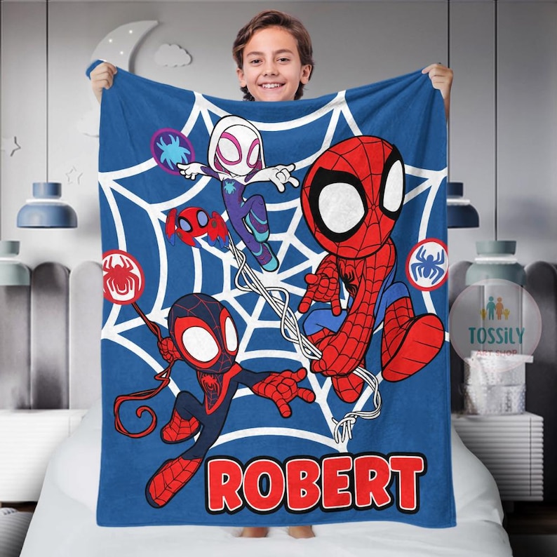 K&ouml;nnte beinhalten: Eine blaue Fleece-Decke mit einem Spinnennetz-Design, das Spider-Man, Spider-Gwen und Miles Morales zeigt. Die Decke tr&auml;gt den Namen "ROBERT" in roten Buchstaben.