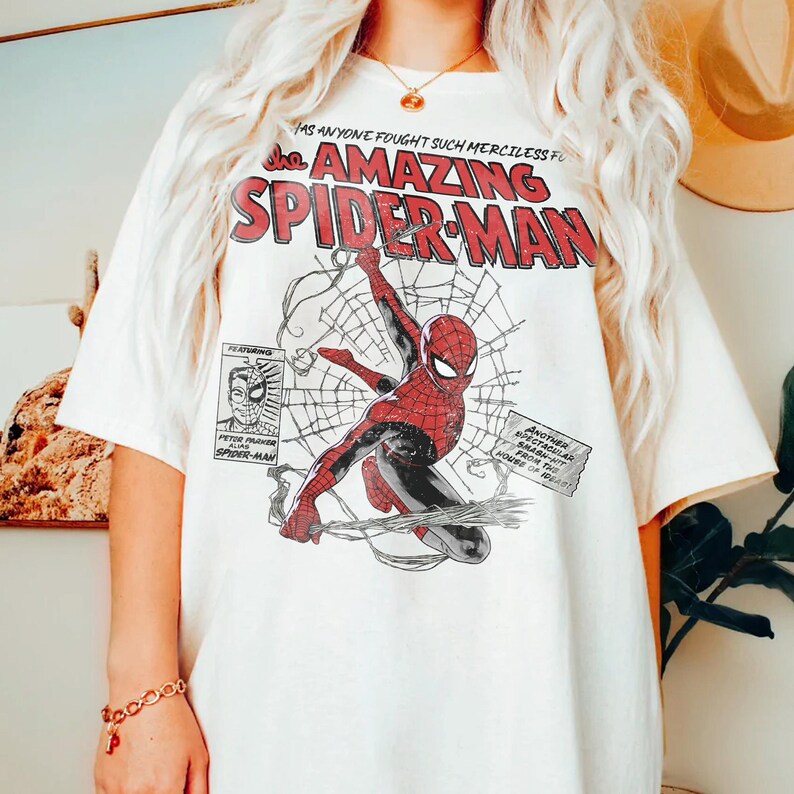 Pu&ograve; includere: T-shirt bianca con una grafica in stile fumetto di Spider-Man in rosso e nero, con la scritta "The Amazing Spider-Man" in rosso. Sono visibili anche altri fumetti e testo.