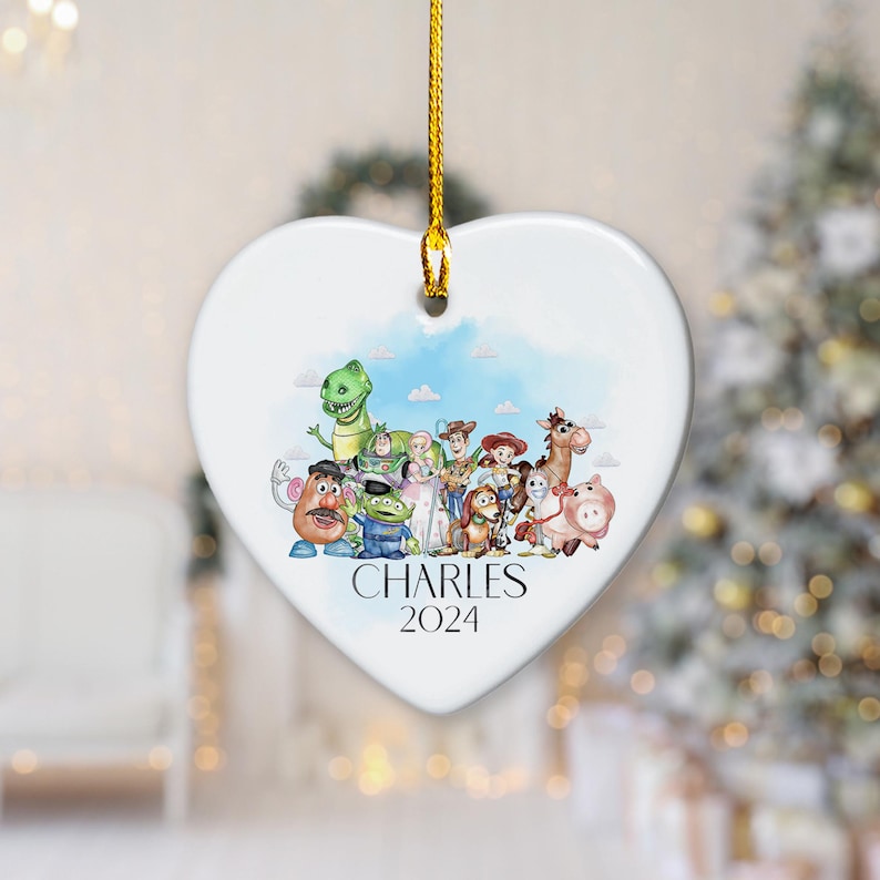 May include: White heart-shaped ceramic ornament featuring a watercolour illustration of characters from the Toy Story film franchise. The ornament has a gold string for hanging and the text "CHARLES 2024" printed on the bottom.
