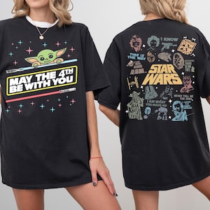 K&ouml;nnte beinhalten: Schwarzes T-Shirt mit einem Baby Yoda-Motiv, Lichtschwertern und dem Text "MAY THE 4TH BE WITH YOU". Auf der R&uuml;ckseite des Shirts befinden sich Illustrationen und Zitate von Star Wars-Charakteren. L&auml;ssiger, &uuml;bergro&szlig;er Schnitt.