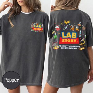 May include: Pepper-colored t-shirt with the text "LAB WEEK" on the front and "LAB STORY" on the back, along with science-themed graphics. The shirt has a crew neck and short sleeves.