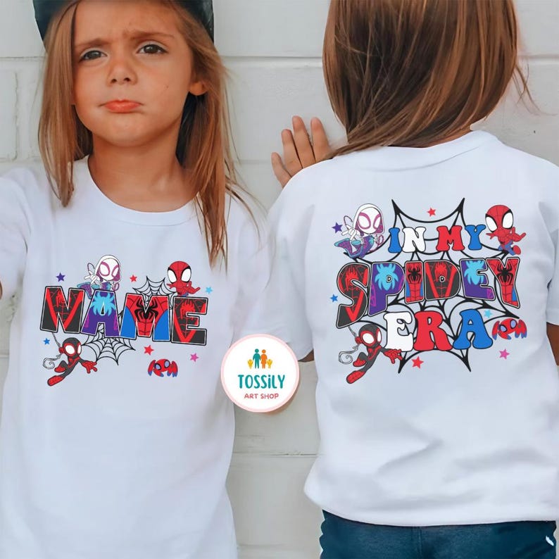 K&ouml;nnte beinhalten: Wei&szlig;e T-Shirts mit farbenfrohen Spider-Man-Grafiken. Ein Shirt zeigt das Wort "NAME" in einem Web-Design, und das andere sagt "IN MY SPIDEY ERA". Beide Shirts haben Spider-Man-Charaktere und Web-Designs.