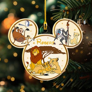 May include: A white, gold, and brown Christmas ornament shaped like Mickey Mouse's head. The ornament features characters from The Lion King, including Simba, Nala, and Mufasa. The ornament is personalized with the name "Robert".