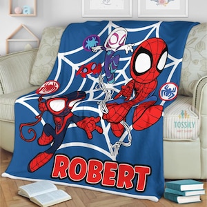 K&ouml;nnte beinhalten: Eine blaue Fleece-Decke mit einem wei&szlig;en Spinnennetz-Design. Die Decke zeigt vier Comic-Spider-Man-Charaktere, darunter Spider-Man, Spider-Gwen und Miles Morales. Die Decke ist mit dem Namen "ROBERT" personalisiert.