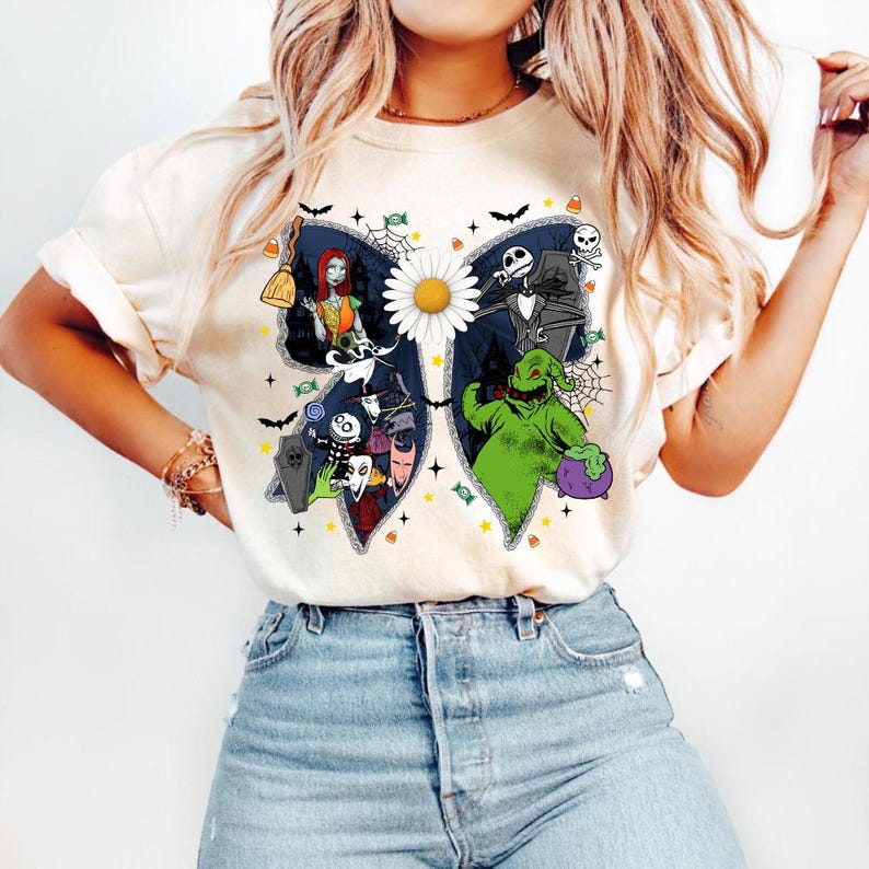 May include: Cream-coloured t-shirt featuring a butterfly-shaped graphic with characters from "The Nightmare Before Christmas." The design includes Sally, Jack Skellington, Oogie Boogie, and other characters, with a daisy in the centre. Halloween-themed.