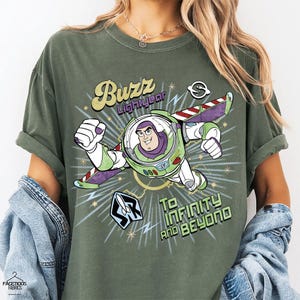 Puede incluir: Camiseta verde oliva con un gr&aacute;fico de Buzz Lightyear en vuelo, con el texto "Buzz Lightyear" arriba y "To Infinity and Beyond" abajo. El dise&ntilde;o incluye acentos verdes, morados, blancos y amarillos.