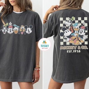 May include: Dark gray t-shirt featuring a classic cartoon character design. The front displays a row of characters, while the back showcases a checkered pattern with more characters and the text "Mickey & Co. EST.1928".