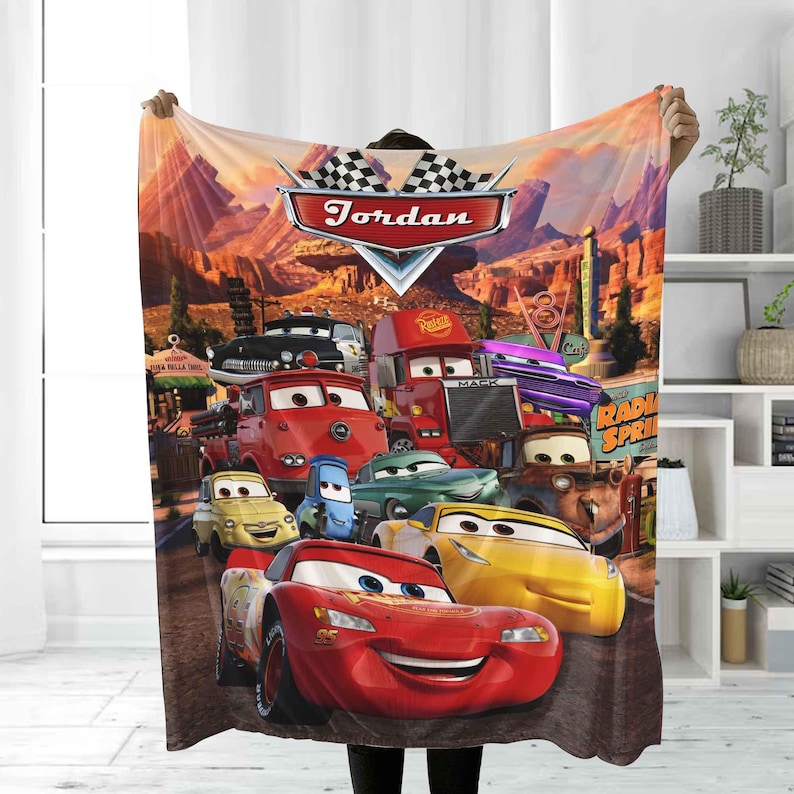 May include: A fleece blanket featuring a colorful design of characters from the animated movie "Cars". The blanket showcases Lightning McQueen and other vehicles in a desert landscape, with the name "Jordan" displayed prominently.